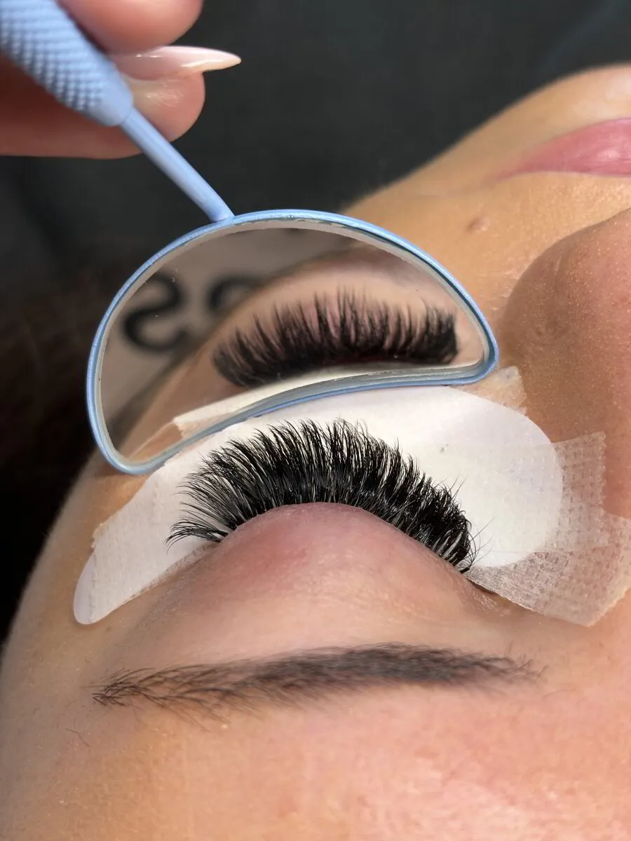 Mary J holding a small mirror over a client's closed eye, showing a full set of volume lashes